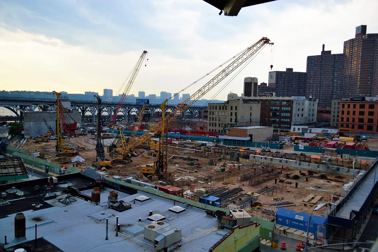 construction site, harlem, new york, uptown, upper west side, residential, buildings, apartments, high rise, foundation, cement, pillars, beams, crane, dump truck, trucks, soil, sand, hard hat, tools, concrete, workers, sky, clouds, rooftop, roof, looking over, high ground, overhead, areal, bridge, train station, riverside drive, river, hudson river, manhattan, gentrification, industrial, landmark, excavation, new, cityscape, riverside, building, architecture, america, usa, day, urban, business, commercial, financial, design, construction, people, scene, tower, real-estate, money, finance, insurance, liability, insured, day time, power, creative, constructor, foreman, demolition, dirty job, discovery, discover, work, hard work, professional, sign, idea, entrepreneur, blue sky, hazard, cones, barriers, blue, brown, nature, beige. Why do a New York City contractor and a rural Texas contractor pay different premiums?