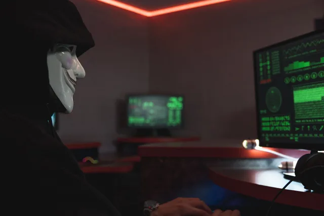 A person wearing a hacker mask operates a computer in a dimly lit room with digital displays. Cyber insurance splits into two halves most buyers never separate